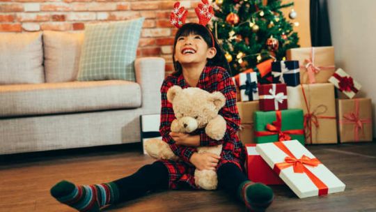 A girl sitting on the ground hugging a teddy with a Christmas tree in the background.