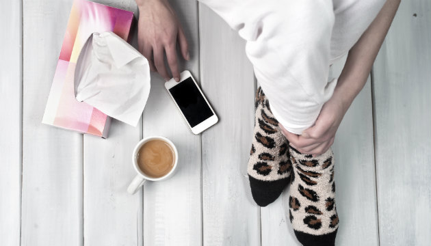 An image of a person with socks on sitting on the ground with a phone, cup of coffee and a box of tissues. | Canstar