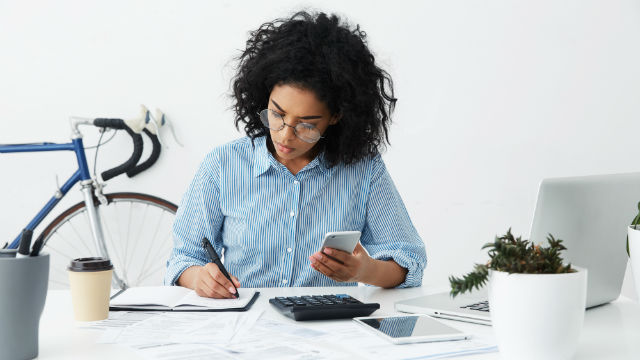 A woman looking at her phone and writing in a journal with a calculator on the desk and a bike in the background.