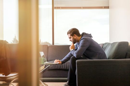 A man is sitting on a lounge across from a laptop and looks like he is thinking