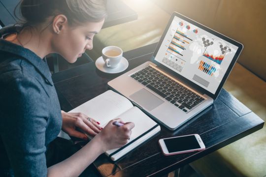 A woman looking at investment graphs on a laptop and writing in a notebook