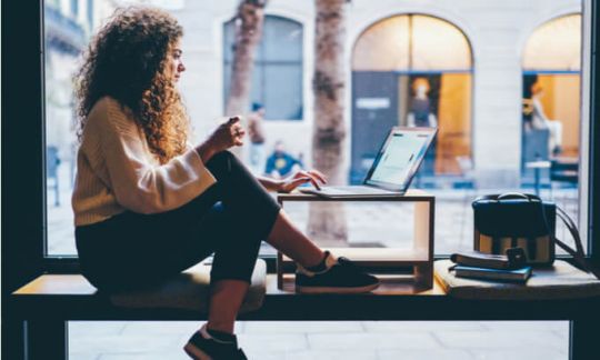 Woman sits on bench working on a laptop drinking a coffee