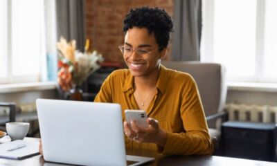 Woman holding a phone and looking at her laptop searching for the highest interest savings account.