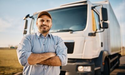 A male truck driver stands in front of his truck with his arms folded.