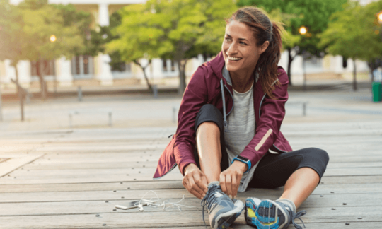 Woman sitting on boardwalk putting on her running shoes, considering private health insurance cost.