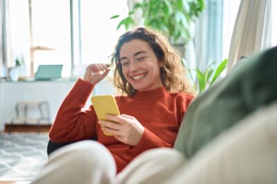 Young woman sitting on a sofa in a light-filled apartment, looking at her smartphone