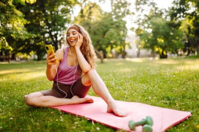 Young woman sitting on yoga mat with her smartphone in a park